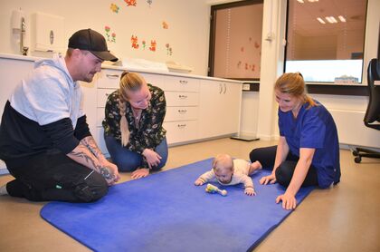 Bildet viser et barn som ligger på gulvet på en matte. Sykepleier sitter på gulvet sammen med mor og far.