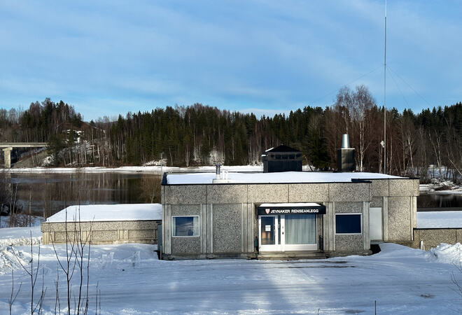 Bildet viser en grå bygning omkranset av snø og fjord. Omtrent midt på bildet er det et inngangsparti, over det står det "Jevnaker renseanlegg".