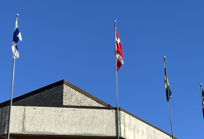 Bildet viser deler av et hustak, og fire flaggstenger som alle har hvert sitt flagg heist. Flaggene henger relativt rett ned grunnet fraværet av vind.