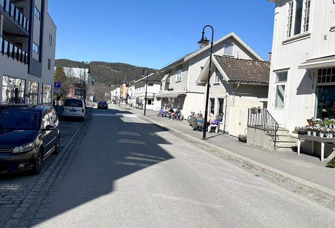 Bildet viser Storgata, fotografen står ute i veien. Til venstre i bildet ser man noen biler stå parkert. Til høyre ser man flere hus, og at det sitter folk på fortauet i sola.