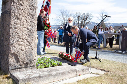 Bildet viser en mann som legger ned en blomsterkrans foran en minnestein. I bakgrunnen ser man en gruppe mennesker som følger med.