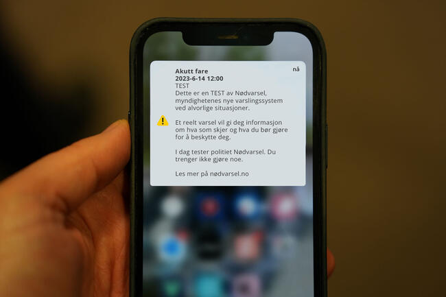 Bildet viser en del av en mobiltelefon, og en del av en hånd som holder denne. På skjermen ses et eksempel på nødvarsel. Teksten sier at det er en test.