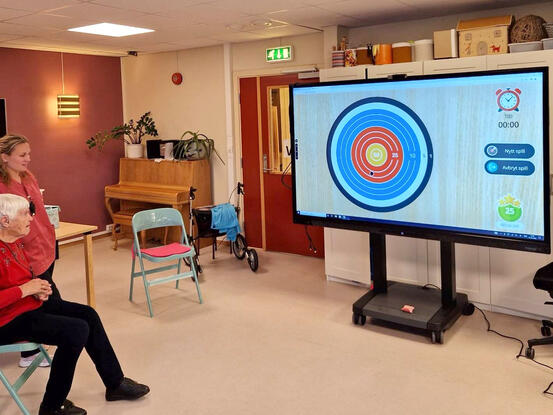 Bildet viser en sittende, eldre kvinne som ser på en stor skjerm med et interaktivt dart-lignende spill. En ansatt står ved siden av henne og følger med.