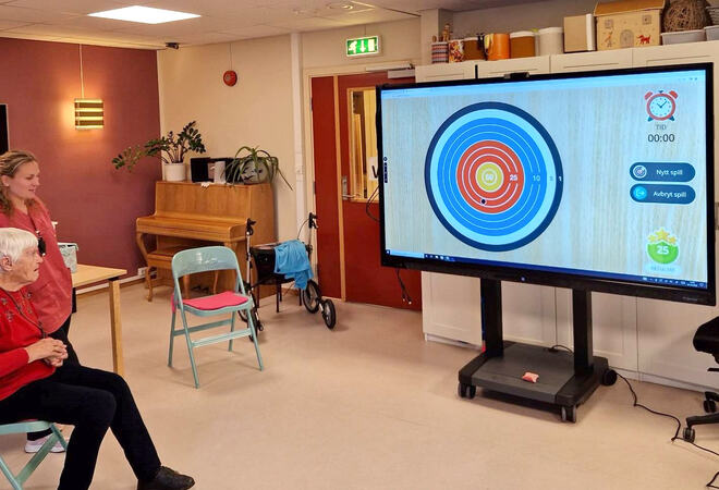 Bildet viser en sittende, eldre kvinne som ser på en stor skjerm med et interaktivt dart-lignende spill. En ansatt står ved siden av henne og følger med.