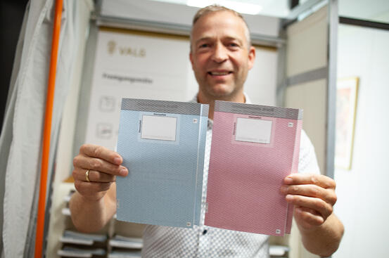 Bildet viser en mann som viser fram to stemmesedler – en blå og en rosa. Han står foran et valgavlukke.