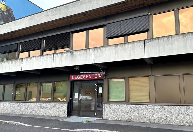 Bildet viser inngangspartiet på Samfunnshuset legekontor. Det er vindusrekker i to etasjer. Omtrent midt på bildet er det en bred dør, med et rødt skilt over. Der står det «legesenter» med store bokstaver.