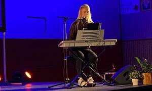 Bildet viser en artist som sitter bak et el-piano. En mikrofon viser at personen også synger. Hun sitter på en scene.