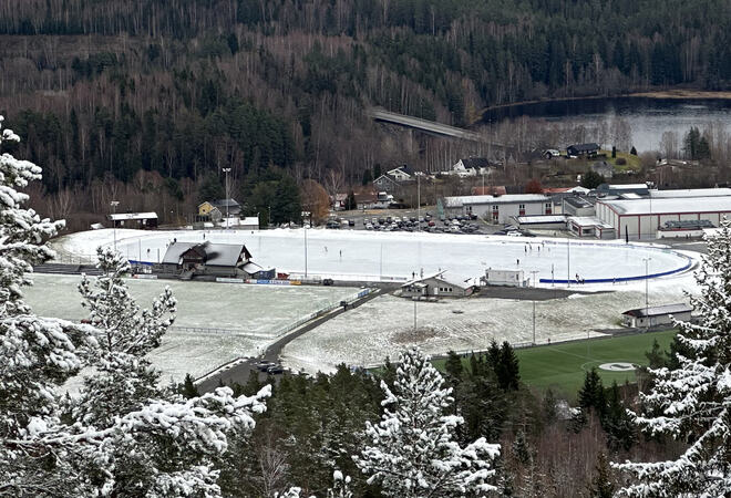 Bildet viser kunstisbanen, det er tatt på lang avstand. Man kan se at det er aktivitet på isen. I forkant ser man gressflater med litt snø på, bak ser man skolen, skog og vann.