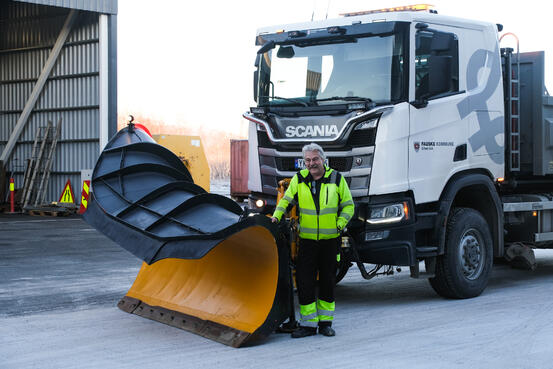 mann i grønn arbeidsjakke står foran lastebil med snøplog