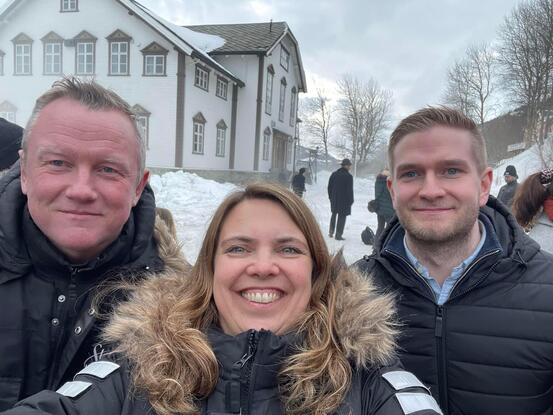 to menn med en kvinne i midten tar selfie og smiler. De er ute med snø og et bygg i bakgrunnen