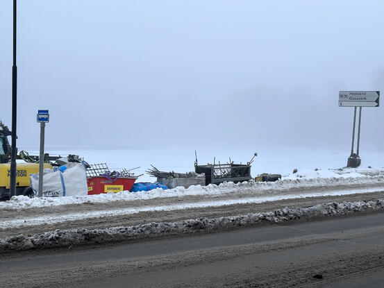Bildet viser en veiskulder dekket i snø. Nærmest ser man bar vei. På skulderen ser man buss-skilt, diverse anleggsutstyr og et veiskilt som peker mot Hadeland Glassverk.