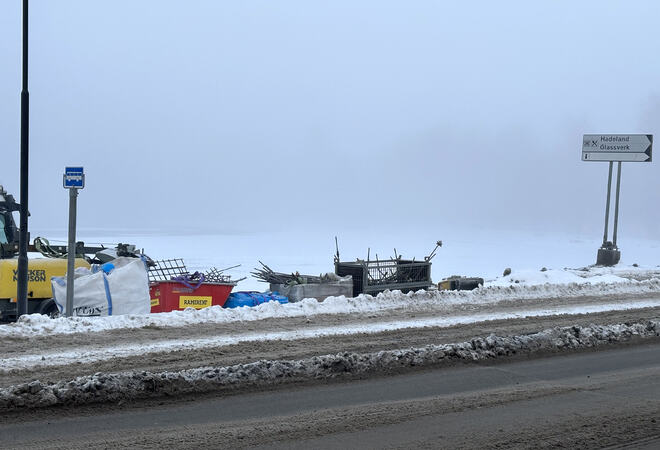 Bildet viser en veiskulder dekket i snø. Nærmest ser man bar vei. På skulderen ser man buss-skilt, diverse anleggsutstyr og et veiskilt som peker mot Hadeland Glassverk.