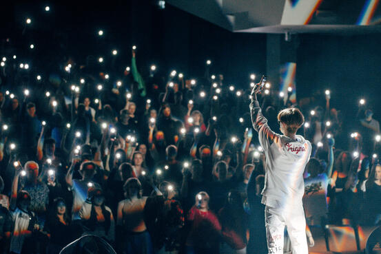 Mann i hvitt står på scene foran publikum som lyser opp med lommelykt på mobil.