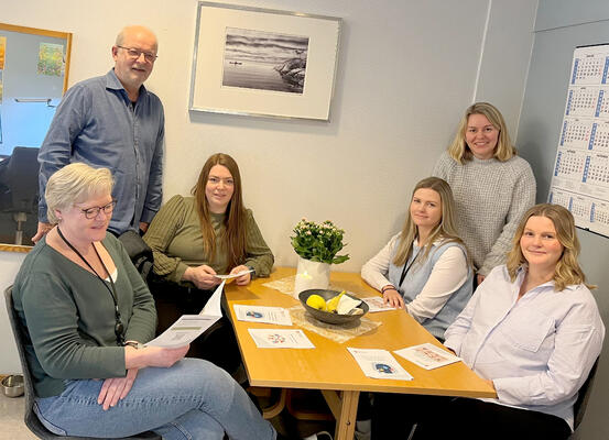 Bildet viser seks personer rundt et bord. To står, de andre sitter. Personen framme til venstre ser i en brosjyre. De andre ser i kamera.