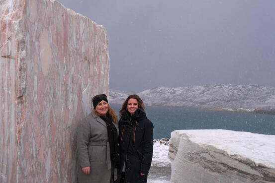 To kvinner står ute foran en marmorinstallasjon. Begge smiler. Sjø og fjell i bakgrunn.