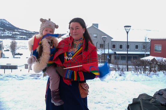En kvinne i kofte smiler og holder et barn på armen. Begge holder samisk flagg. De er ute med snø og bygg i bakgrunnen