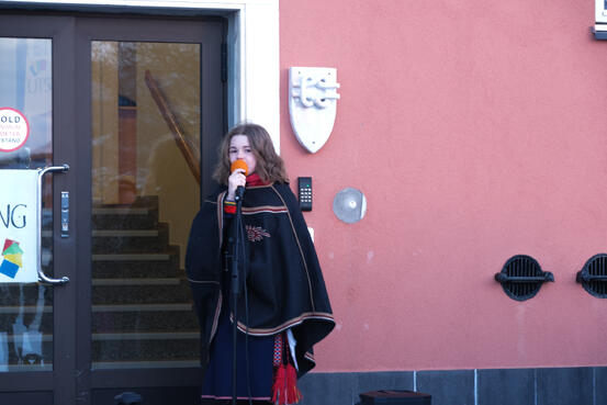 Jente i samiske klær synger i mikrofon. Hun står utenfor et bygg med en knute laget av marmor på veggen
