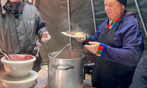 To menn inne i en lavvo som står foran store gryter og serverer mat.