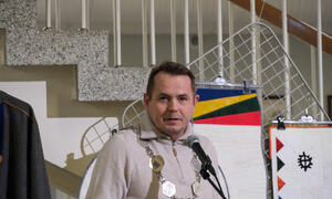Mann holder tale med samisk brukskunst i bakgrunn. Han har på seg ordførerkjede