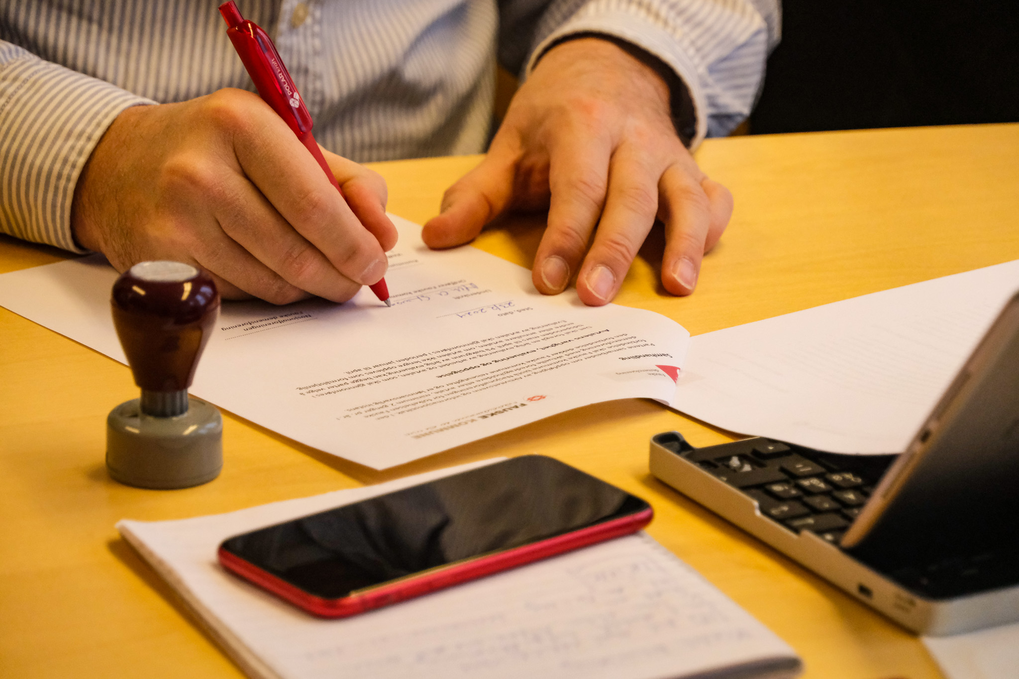 Signering av avtale med rød penn. Et stempel står på bordet ved siden av. En telefon ligger foran