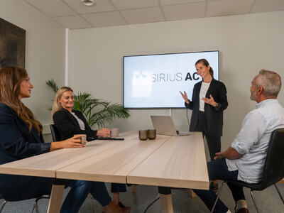 Bildet viser fire personer rundt et bord. En av dem står, to av de andre ser på denne. Det er et stort møtebord uten rot på. På veggen henger en skjerm hvor det står "Sirius Act".