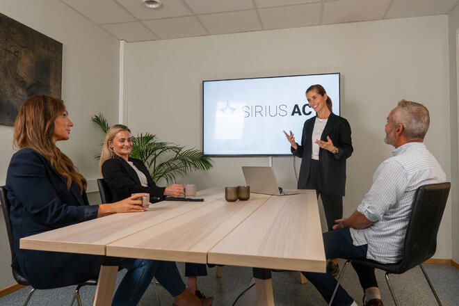 Bildet viser fire personer rundt et bord. En av dem står, to av de andre ser på denne. Det er et stort møtebord uten rot på. På veggen henger en skjerm hvor det står "Sirius Act".