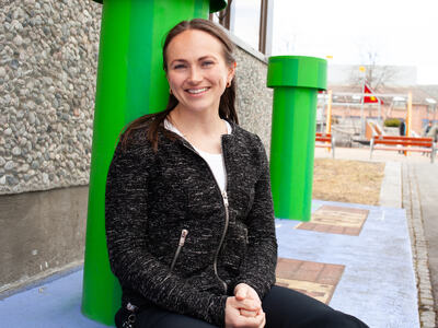 Bildet viser en person som sitter på en malt betongflate. Hun støtter seg på en grønn søyla. I bakgrunnen ser man en til lik søyle. Man ser veggen på en bygning, med grov steinvegg. I bakgrunnen ses en fargerik lekeplass. Personen har langt hår so
