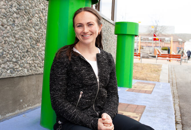 Bildet viser en person som sitter på en malt betongflate. Hun støtter seg på en grønn søyla. I bakgrunnen ser man en til lik søyle. Man ser veggen på en bygning, med grov steinvegg. I bakgrunnen ses en fargerik lekeplass. Personen har langt hår so