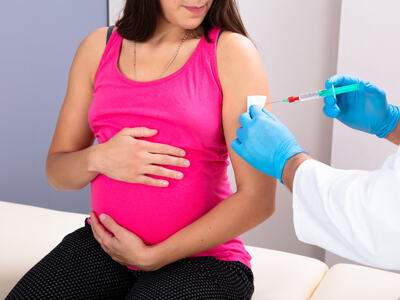Bildet viser en gravid sittende på en benk. Hun holder rundt magen sin og ser ned til sin venstre. Der settes det en vaksine. Vi ser kun hendene til vedkommende, som har hvit frakk og blå hansker.