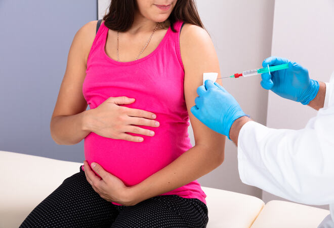 Bildet viser en gravid sittende på en benk. Hun holder rundt magen sin og ser ned til sin venstre. Der settes det en vaksine. Vi ser kun hendene til vedkommende, som har hvit frakk og blå hansker.