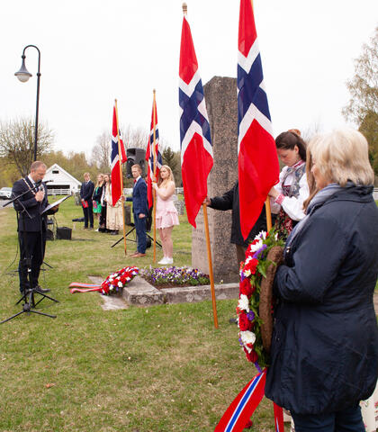 Bildet viser en mann som står foran en bauta. På bakken ligger en blomsterkrans. Fire personer holder fire, norske flagg. Nærmest står en person med ytterligere en blomsterkrans. Bildet er tatt fra siden av de som står.