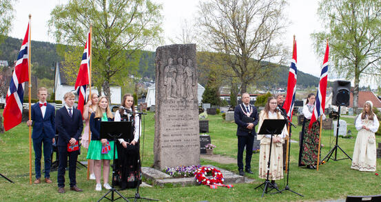Bildet viser totalt ti personer som står på hver side av en stor bauta. Fire av dem står med store norske flagg.