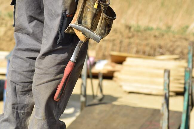 Bildet viser et slitent verktøybelte med en hammer hengende ved to bein. I bakgrunnen ser man planker og stålrør.