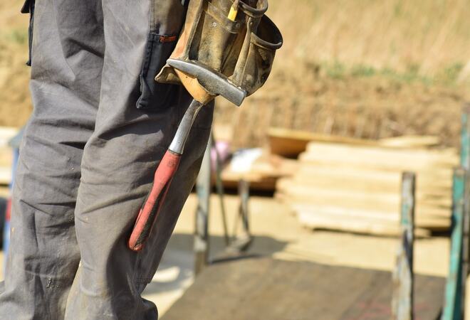 Bildet viser et slitent verktøybelte med en hammer hengende ved to bein. I bakgrunnen ser man planker og stålrør.
