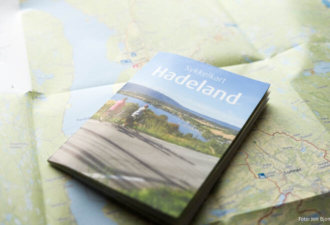 Bildet viser et sammenbrettet kart med bilde av to syklister som ser utover Randsfjorden. På papiret står det «Sykkelkart Hadeland». Det ligger oppå et utbrettet kart.