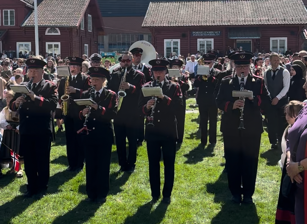 Bildet viser mennesker som spiller i korps. Bildet viser korps i uniform foran rød bygning
