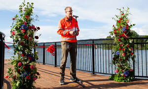 Bildet viser en anleggskledd mann som står mellom to store blomsteroppsatser. I bakgrunnen ser man Randsfjorden.