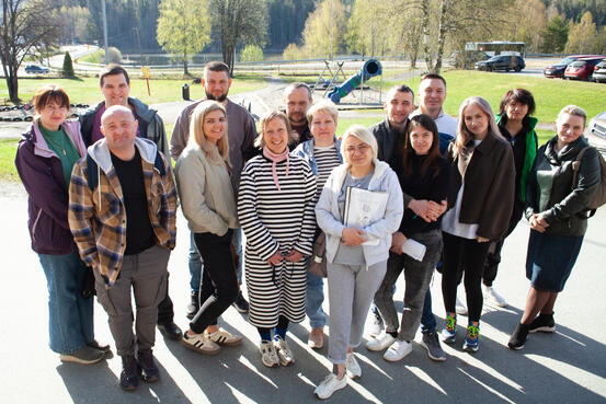 Bildet viser en gruppe totalt 15 personer. De ser i kamera og smiler. De står utendørs. Det er sol, men påkledningen viser at det ikke er veldig varmt. I bakgrunnen ses en lekeplass og noen parkerte biler.