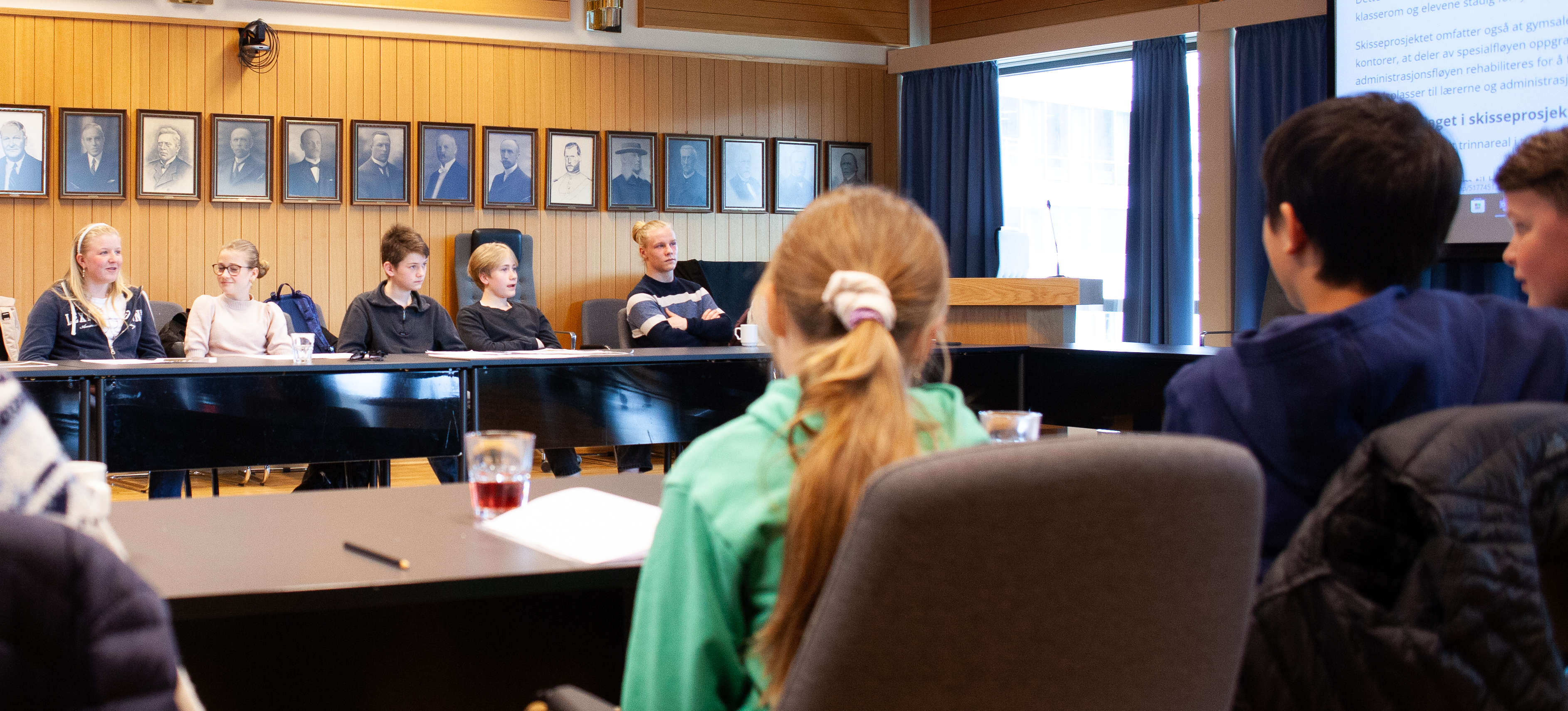 Bildet viser ryggene til et noen elever. Mellom dem ser vi over på den andre bordrekken, hvor det sitter fire elever fra Toso skole samt en representant fra ungdomsrådet.