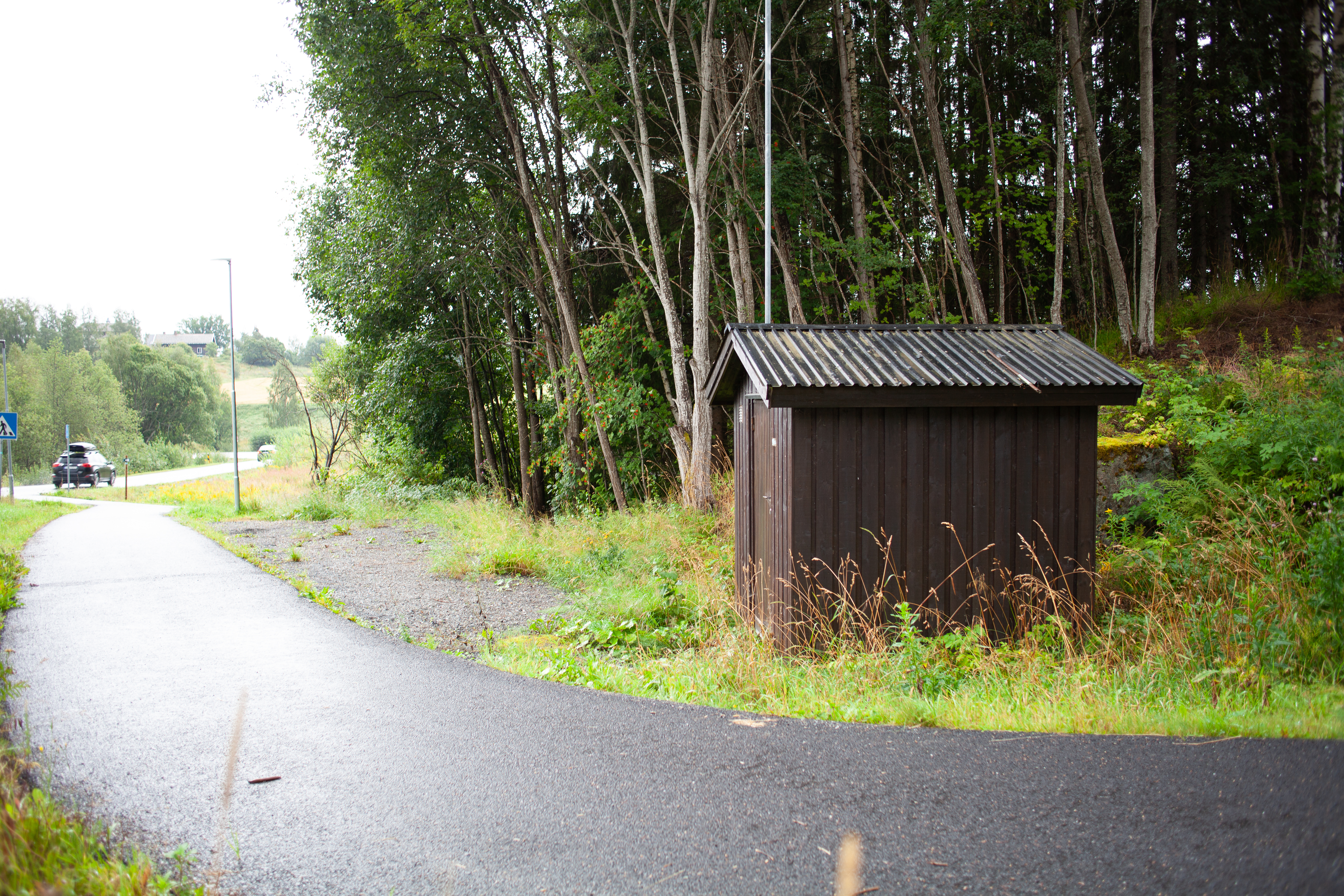 Bildet viser en asfaltert gangvei som går foran en liten bygning. Bak denne er det skog.