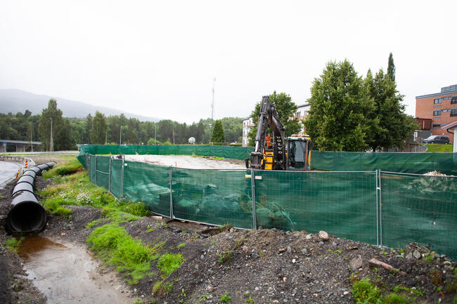 Bildet viser et område med anleggsarbeid. Fra venstre ser man et stort vannrør, og noe vann i forkant. Deretter er det grønt skillegjerde, og bak dette står det en gravemaskin. Bak denne er det noen trær, og man kan se litt av en stor bygning bak dem