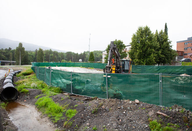 Bildet viser et område med anleggsarbeid. Fra venstre ser man et stort vannrør, og noe vann i forkant. Deretter er det grønt skillegjerde, og bak dette står det en gravemaskin. Bak denne er det noen trær, og man kan se litt av en stor bygning bak dem