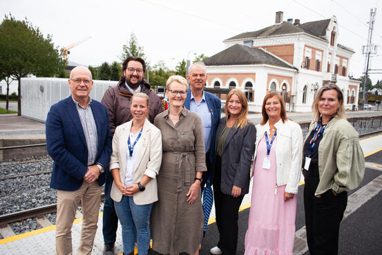 Bildet viser åtte personer som står ved siden av og delvis hverandre. Bak dem ser man en togstasjon.