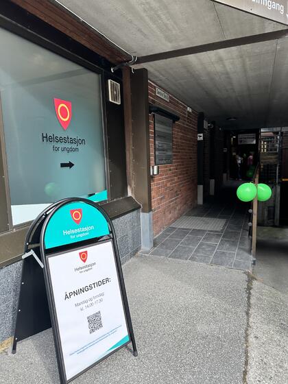 BIldet viser inngangspartiet til helsestasjonen. Det står en plakatbukk med informasjon om åpningstider. På vinduet står det «Helsestasjon for ungdom» og på gelenderet henger det et par ballonger.