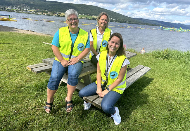 Bildet viser tre personer som sitter på trebenk ved Randsfjorden. De har natteravn-vester på seg, er sommerlig kledt og ser i kamera med smil. I bakgrunnen ser man badeparken.