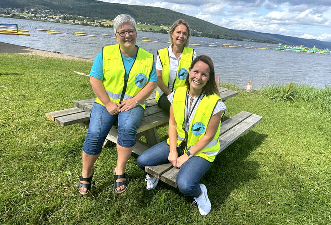 Bildet viser tre personer som sitter på trebenk ved Randsfjorden. De har natteravn-vester på seg, er sommerlig kledt og ser i kamera med smil. I bakgrunnen ser man badeparken.