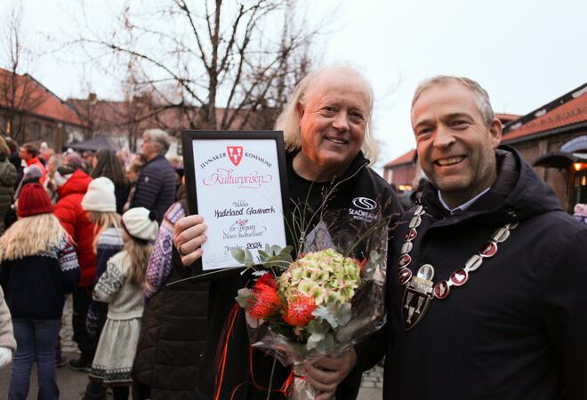 Bildet viser to personer som står ved siden av hverandre. Begge smiler og ser rett i kamera. Den ene holder et diplom og blomster, den andre har ordførerkjede på seg. Bak dem står det masse folk.