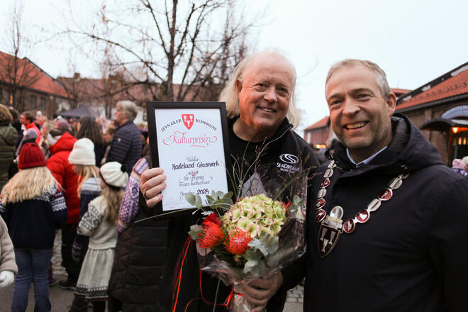 Bildet viser to personer som står ved siden av hverandre. Begge smiler og ser rett i kamera. Den ene holder et diplom og blomster, den andre har ordførerkjede på seg. Bak dem står det masse folk.