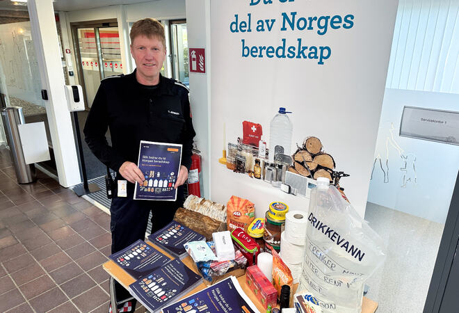 Bildet viser en mann som står bak et bord som er fylt med brosjyrer, matvarer og drikkevann. Det er en stor plakat bak bordet hvor det står «Du er en del av Norges beredskap».