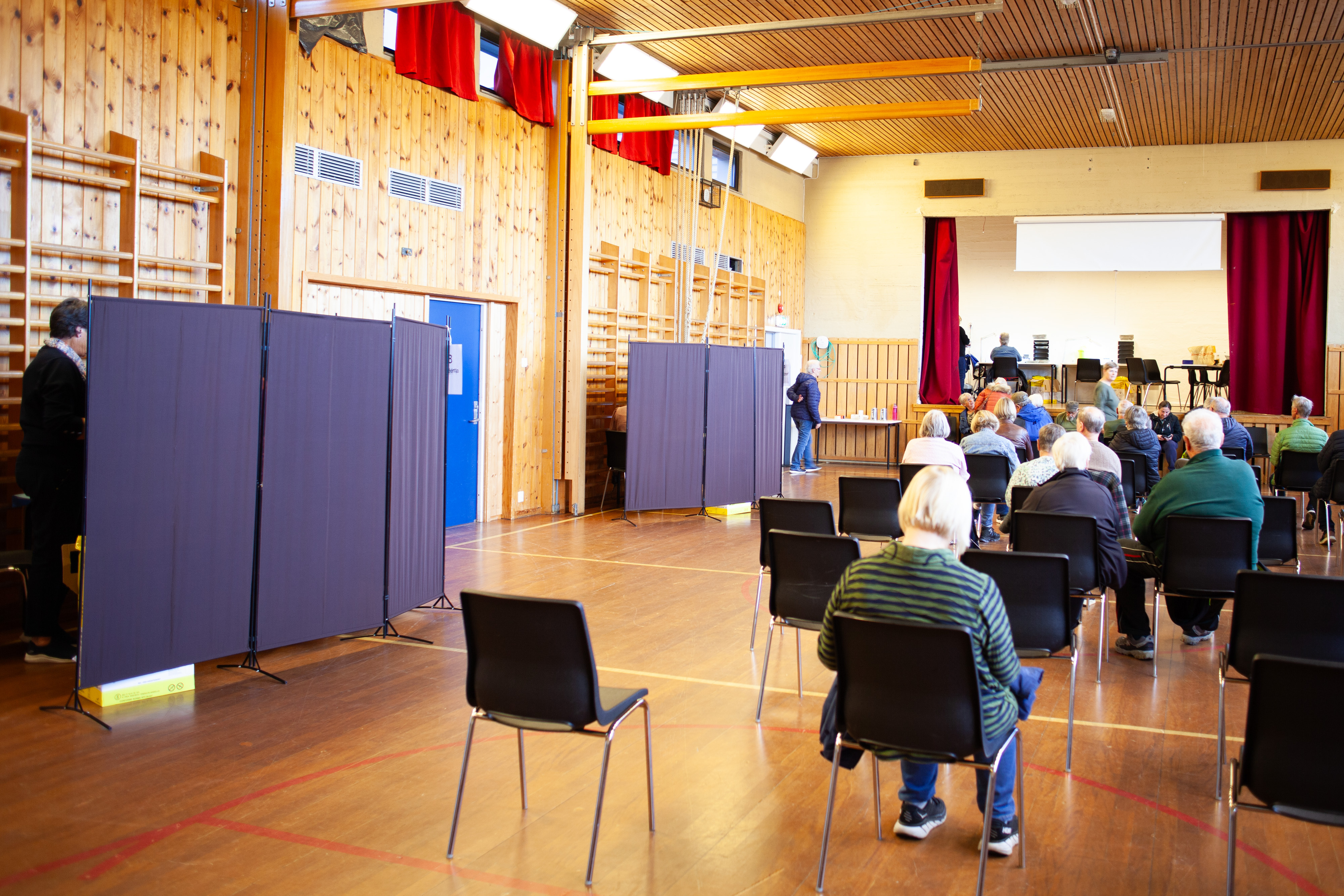 Bildet viser en stor gymsal med båser og personer som sitter på stoler.